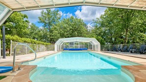 Une piscine à eau claire, protégée par une enceinte en verre et entourée d’une clôture, ainsi qu’un espace de repos couvert avec des chaises.