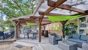 Une terrasse extérieure avec des tables et des chaises en bois, des canapés en osier et une tonnelle verdoyante.