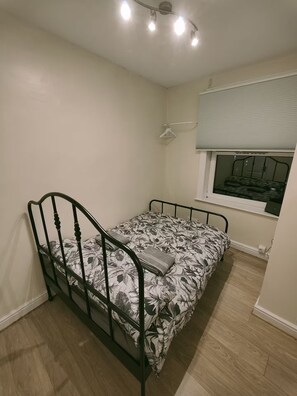 A small, single-bed room with a wooden floor, a window with blinds, and a ceiling light fixture.