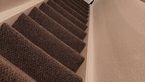 Eine Teppichverkleidete Treppe mit Handlauf, weißen Wänden und einer Leuchte am oberen Ende.