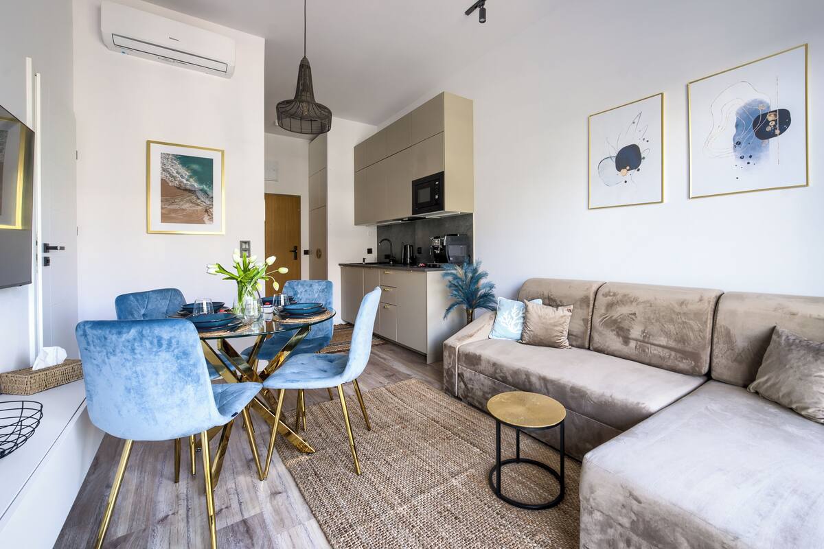 Een moderne woonkamer met een beige bank, blauwe stoelen, een glazen eettafel en een keuken op de achtergrond.