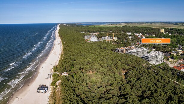 Vista aerea di una zona costiera con spiaggia, fitta foresta e diversi edifici, tra cui un hotel con la scritta "HEVENIA".