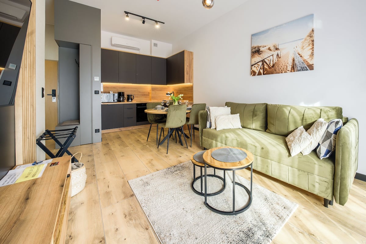 Een moderne woonkamer met een groene bank, een eettafel met stoelen, een keukenhoek en een groot schilderij aan de muur.