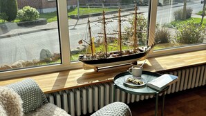 A cozy room with a large window, a model ship, a small table with a cup and saucer, and a cushioned bench. - Vacation apartment no. 2 with its own jetty/barge (Maasholm)