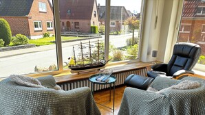 A cozy room with two patterned armchairs, a black leather chair, a small table with a ship model, and a view of a street with houses. - Vacation apartment no. 2 with its own jetty/barge (Maasholm)