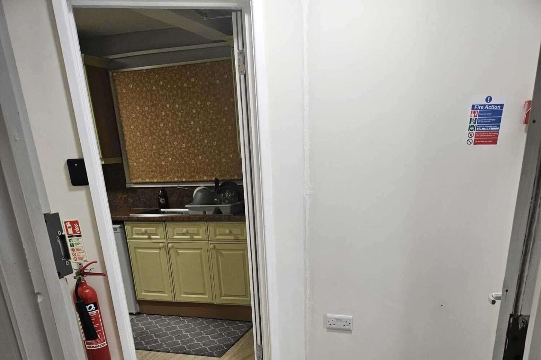 Un pasillo estrecho que conduce a una cocina con un salpicadero decorado, un extintor y un letrero que indica 'Fire Action'.