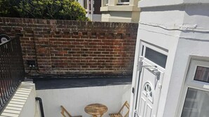 Un pequeño balcón al aire libre con un muro de ladrillo, una puerta blanca y dos sillas de madera.