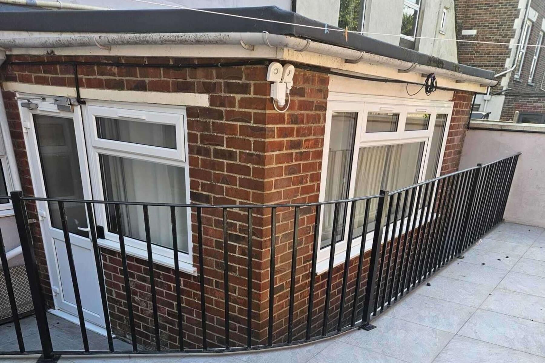 Een klein huis van baksteen met twee ramen, een zwart metalen hekwerk en een betegeld terras.