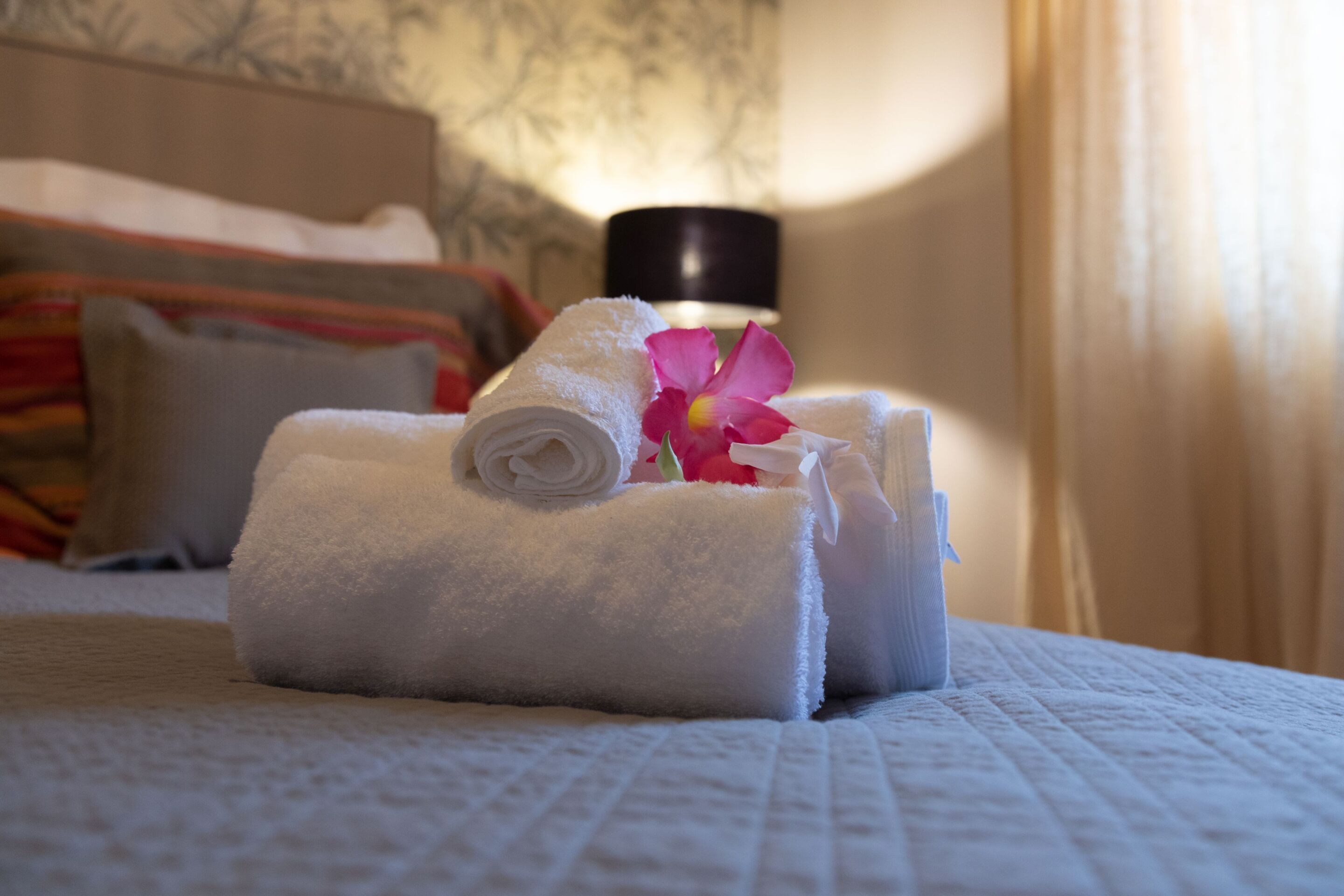 Un letto rifatto con asciugamani bianchi arrotolati e un fiore rosa, una lampada nera e una parete decorata sullo sfondo.