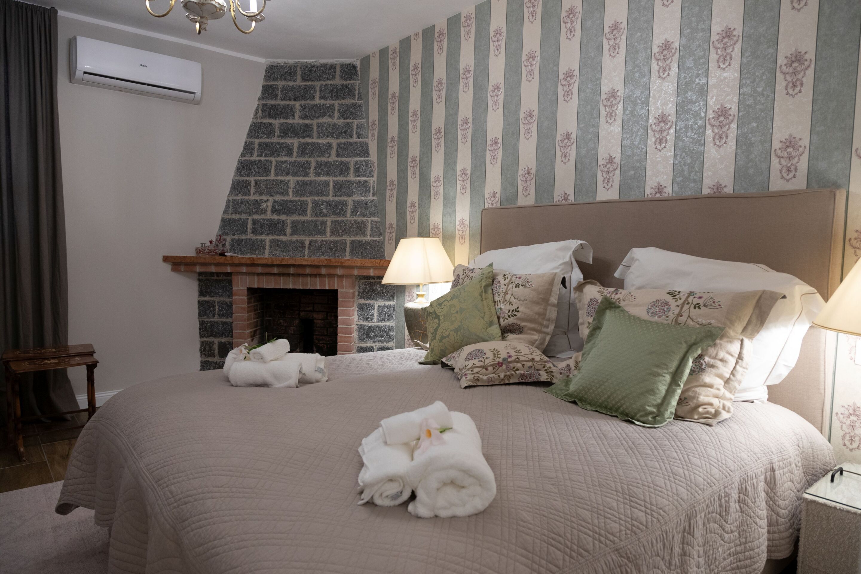 A bedroom with a brick fireplace, a large bed with multiple pillows, and a bedside table with a lamp.