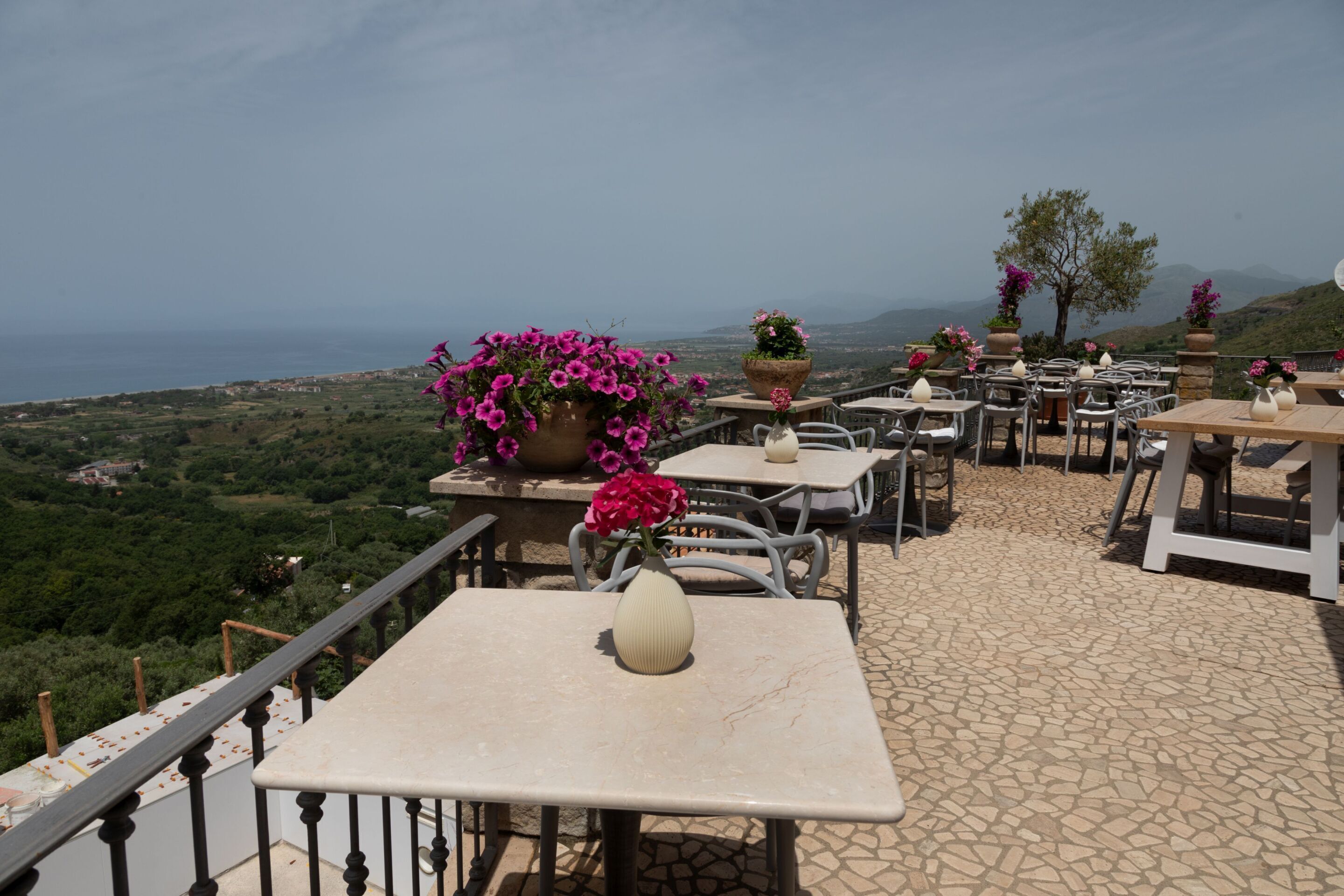 A terrace with tables and chairs, potted plants, and a view of the sea.