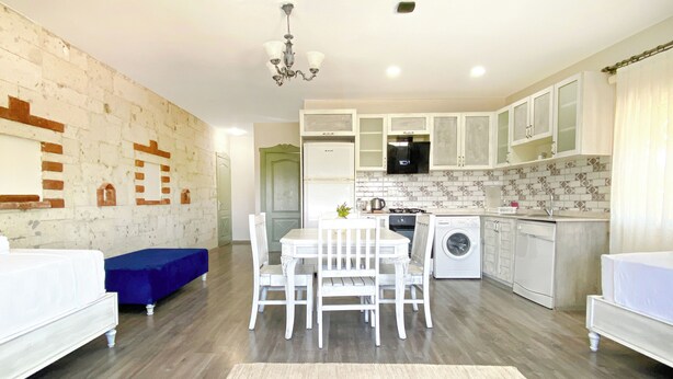 A modern kitchen with white cabinets, a built-in oven, and a microwave. There is a dining area with a white table and chairs, and a blue sofa. - Cumbalıca Flora  (Cesme)