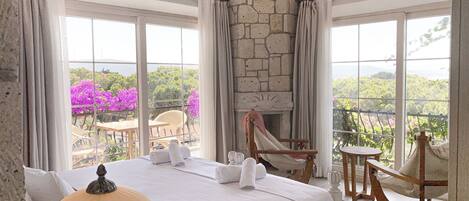 Habitación de hotel con cama, mesa, silla y chimenea. Amplias ventanas ofrecen vistas a árboles y flores.