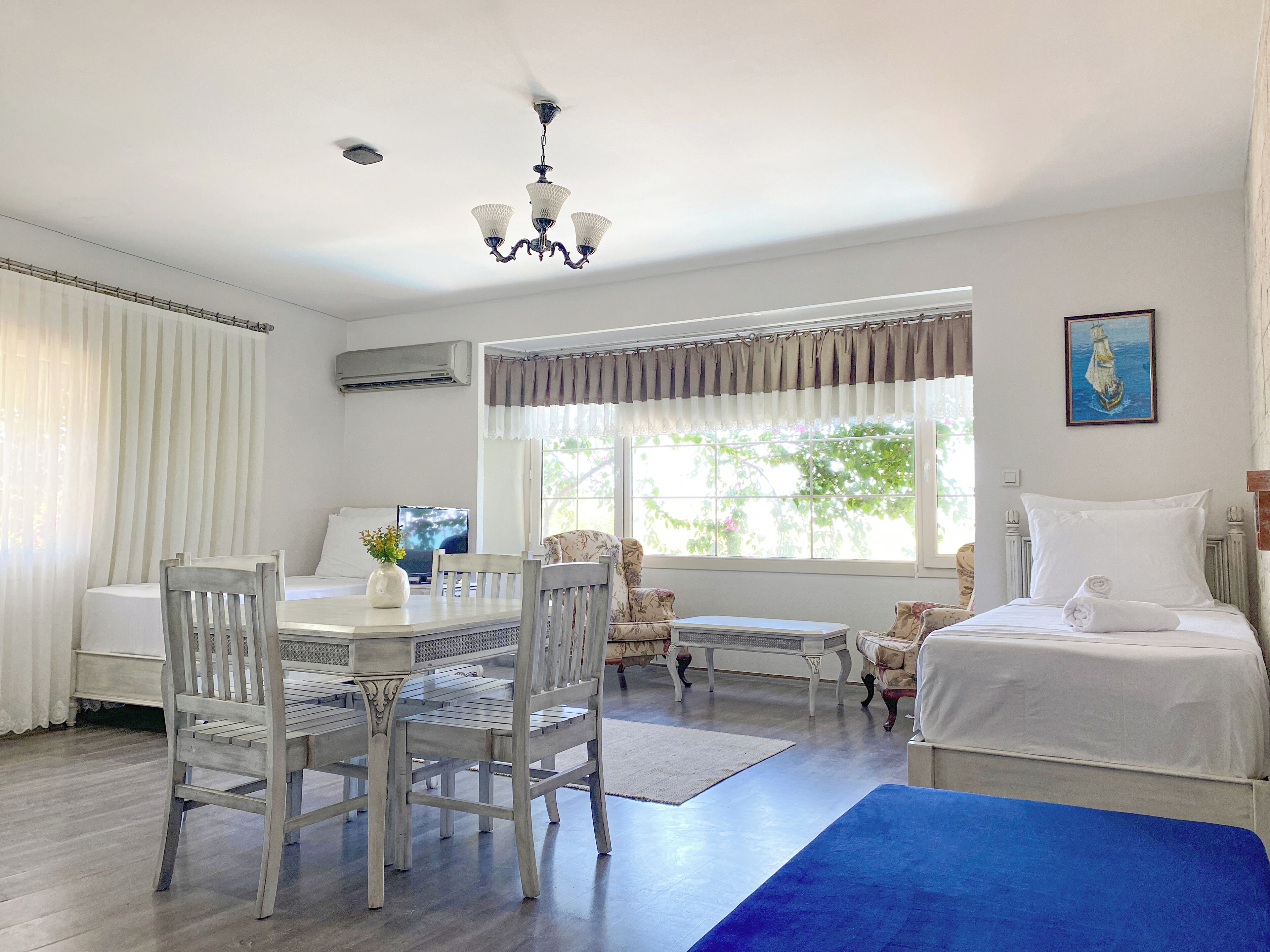 A hotel room with a dining area, a bed with a blue accent, a ceiling fan, and a window with curtains.
