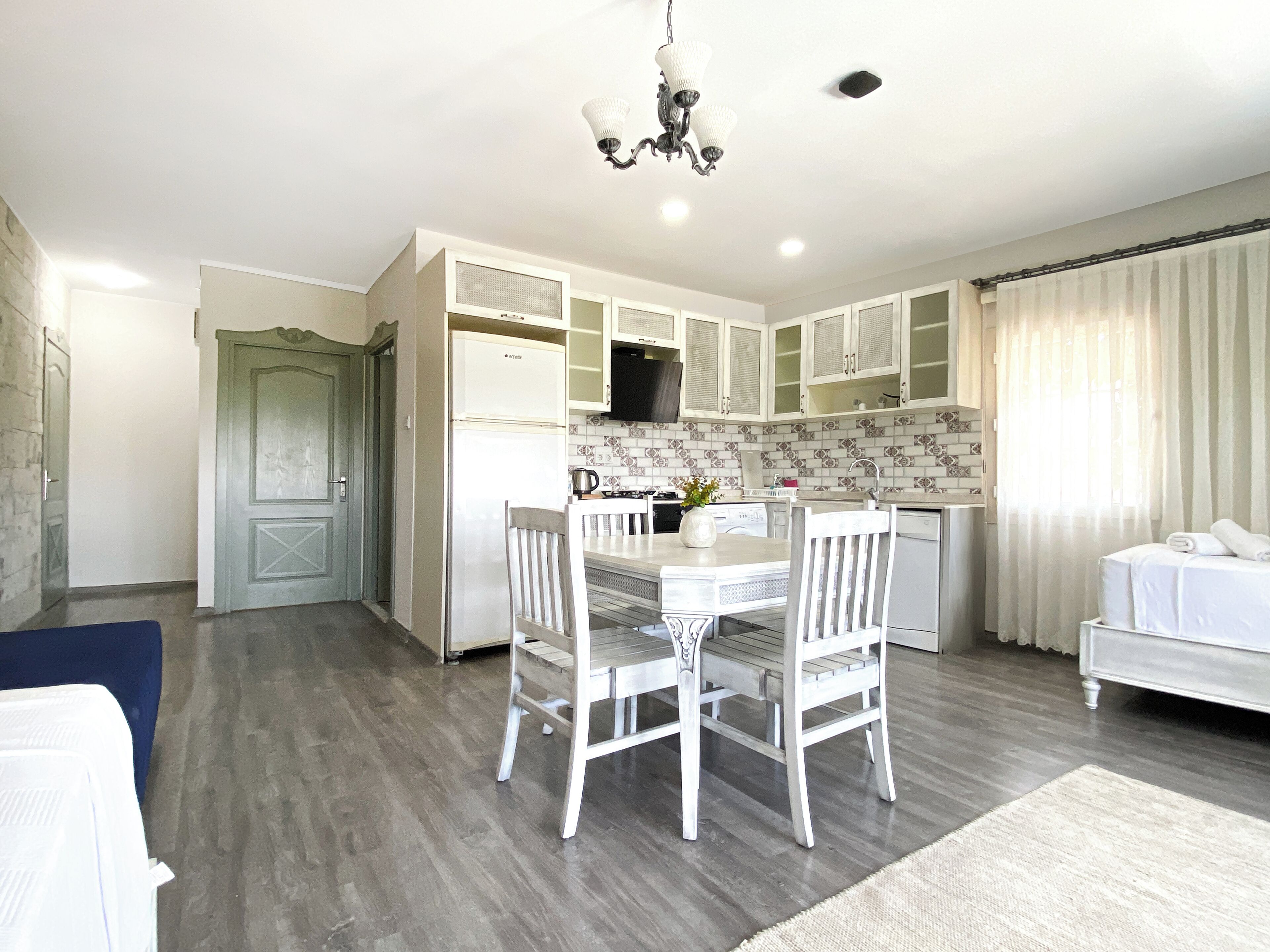A modern kitchen with white cabinets, a dining table with chairs, a chandelier, and a view of the living area with a sofa and curtains.