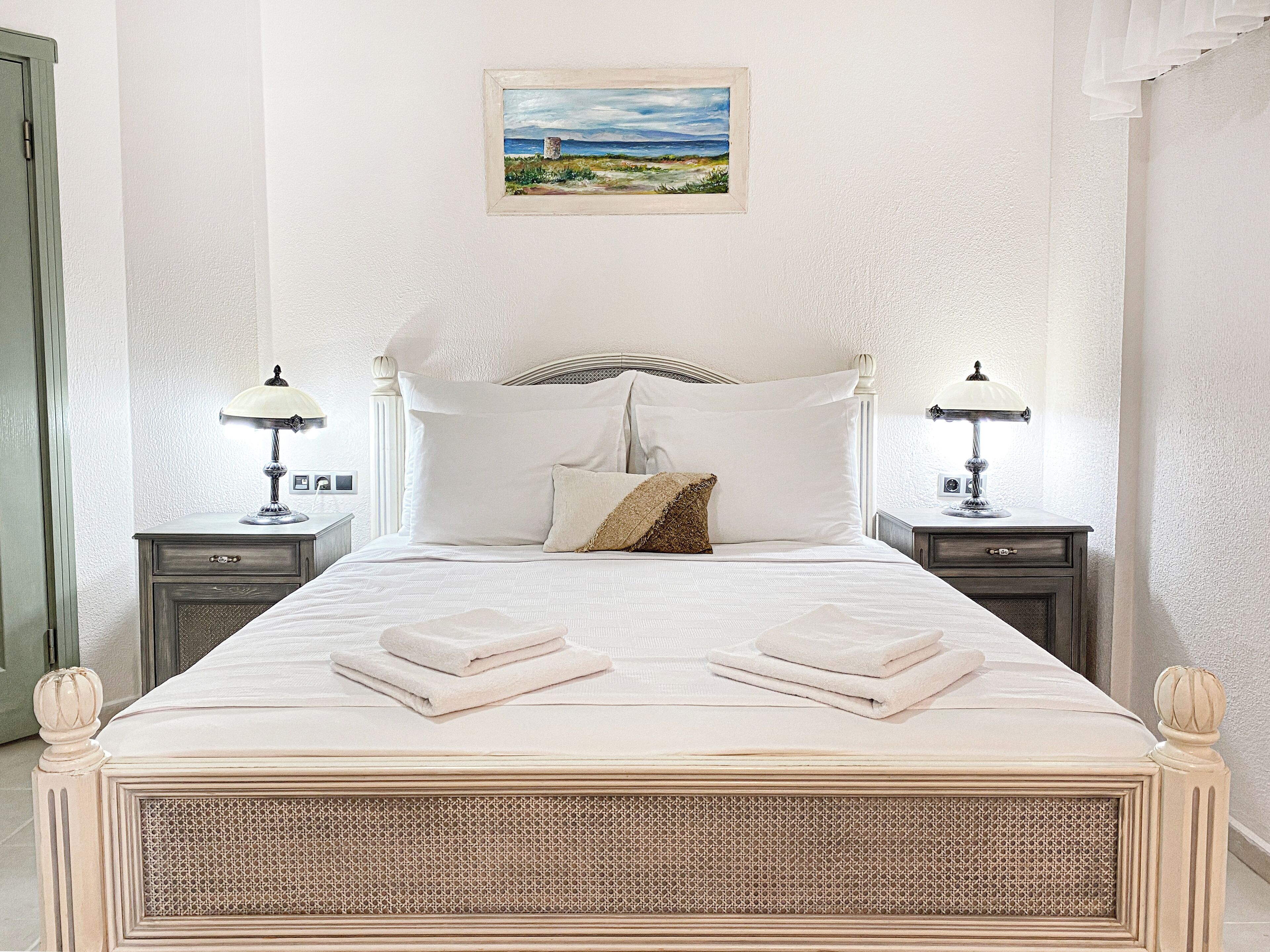 Un letto rifatto con due cuscini e due asciugamani piegati, un quadro raffigurante un paesaggio sulla parete e due lampade da comodino su poggiatesta in legno.
