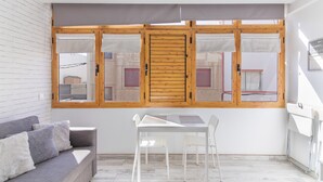 Una habitación con pared de ladrillo blanco, un sofá gris, una mesa blanca con sillas y un ventanal con contraventanas de madera.