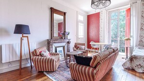 Een woonkamer met gestreepte banken, een open haard, een kroonluchter en een uitzicht door een raam met rode luiken.