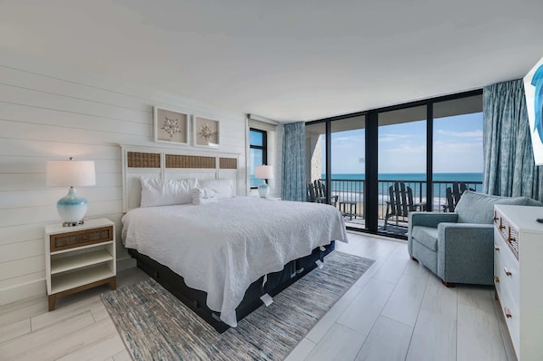 A modern bedroom with a large bed, a nightstand, a dresser, and a view of the ocean.