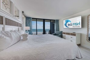 A hotel room with a large bed, a TV, a view of the sea, and a 'Big Fish Rentals' sign.