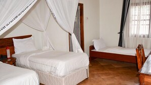 Une chambre à coucher avec deux lits, une tête de lit en bois, une table de chevet sur laquelle se trouve un objet rouge, et une fenêtre avec des rideaux légers.