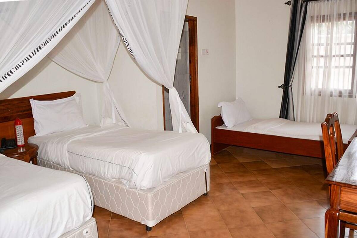 A bedroom with two beds, a wooden headboard, a bedside table with a red object, and a window with sheer curtains.