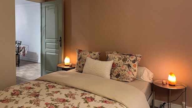 Une chambre à coucher avec un lit, une literie à motifs floraux, deux lampes de chevet et une porte donnant sur une autre pièce.