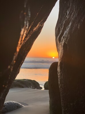 Vista del atardecer a través de una formación rocosa, con una playa y el océano al fondo.