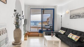 A living room with a black sofa, a white table, a vase with plants, a view of the sea through a glass door, and a painting on the wall. - Stunning Sunset Ocean View Vacation – Ocean Views, Shared Pool & Wi-Fi (Taurito)