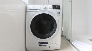 A front-loading washing machine in a bathroom with a glass shower door and a white towel on the floor. - Monthly Rental in Madrid - Modern 1 bedroom flat (Madrid)