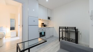 A modern kitchen with white cabinets, a microwave, and a coffee maker. There is a glass table and a gray sofa in the living area. - Monthly Rental in Madrid - Modern 1 bedroom flat (Madrid)