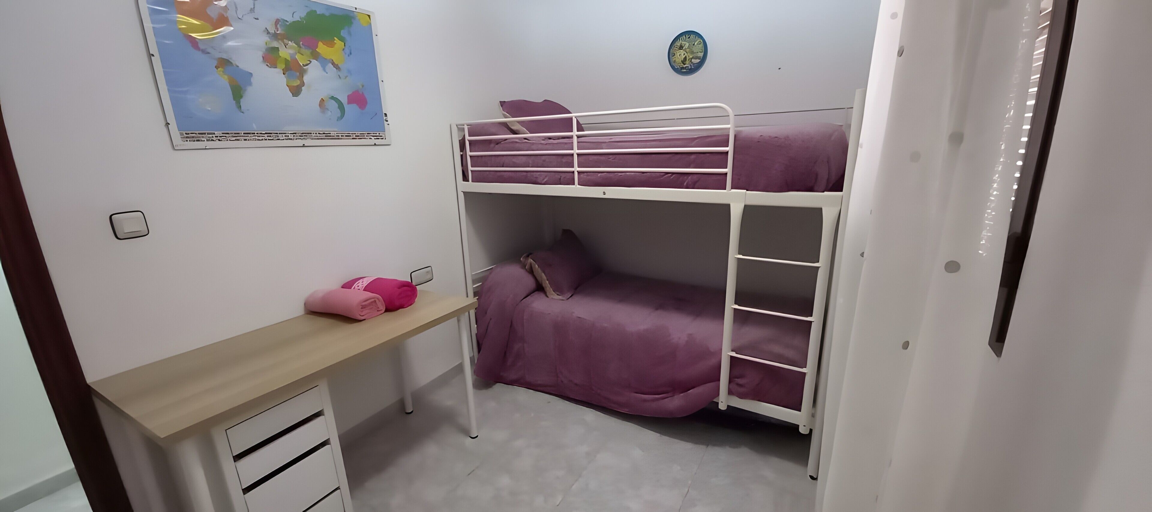 Ein Etagenbett mit violetter Bettwäsche, ein Schreibtisch mit Schubladen, eine Weltkarte an der Wand und ein kleiner, runder Schmuckgegenstand.
