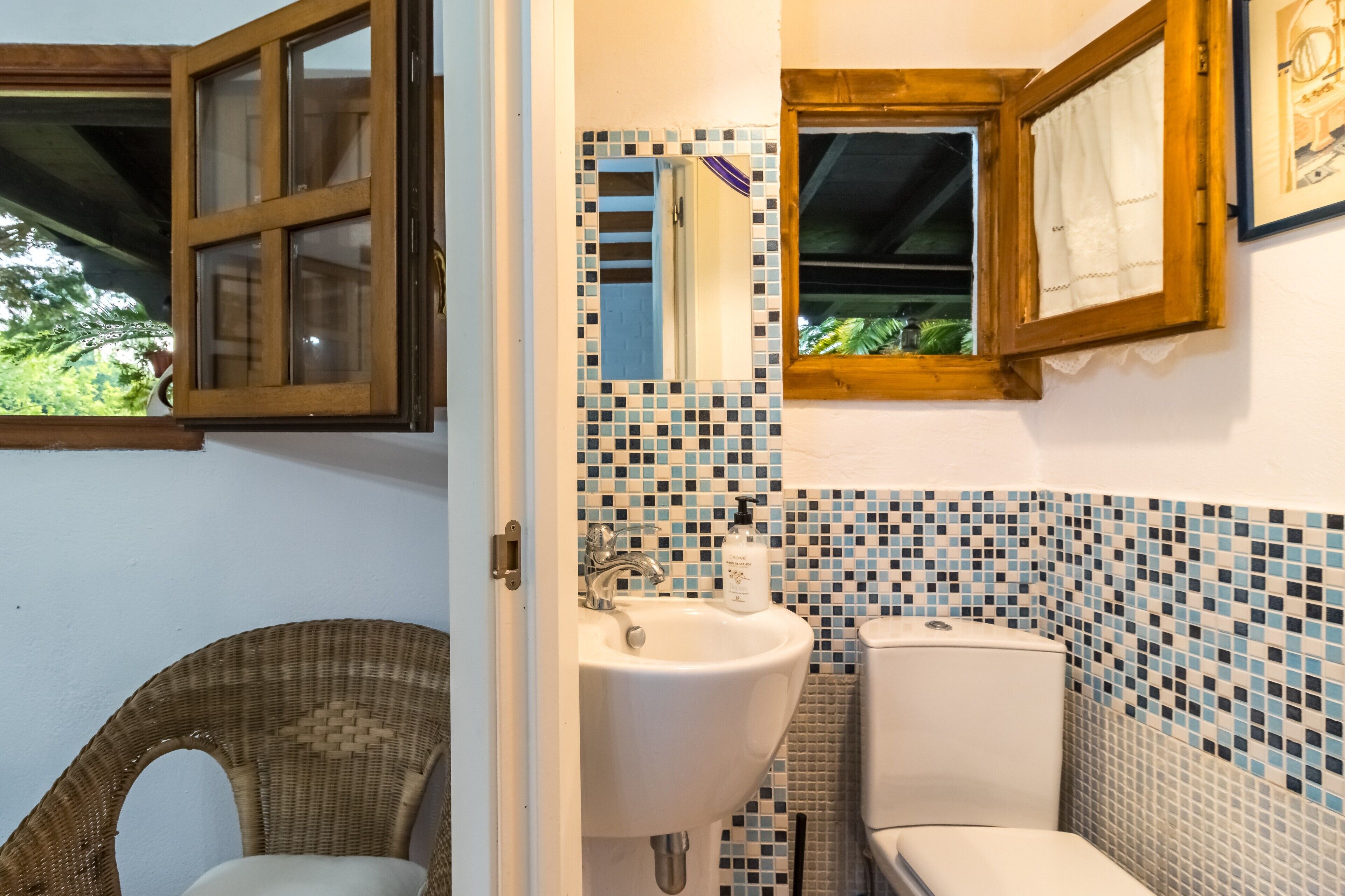 Un baño con una silla de mimbre, un lavamanos, un inodoro y una ventana con cortina.