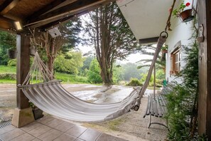 Una cadira espanyola penjant sota una estructura de fusta, amb vista a un jardí i un edifici al fons.