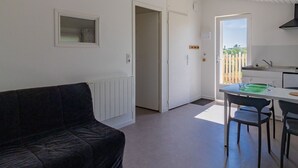 Een compact woongedeelte met een zwarte bank, een eethoek met tafel en stoelen, een kitchenette, en een deur naar een balkon met uitzicht.