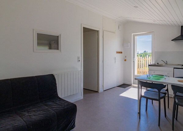 A compact living space with a black sofa, a dining area with a table and chairs, a kitchenette, and a door leading to a balcony with a view. - Cozy studio with terrace in the countryside, near Nantes (Orée-d'Anjou)