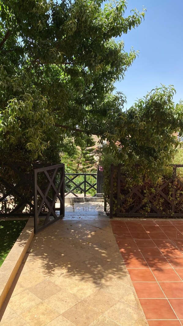 A pathway leading through a garden with a metal gate, surrounded by lush greenery and a tiled floor. - Alwadi Touristic Resort (Taif)