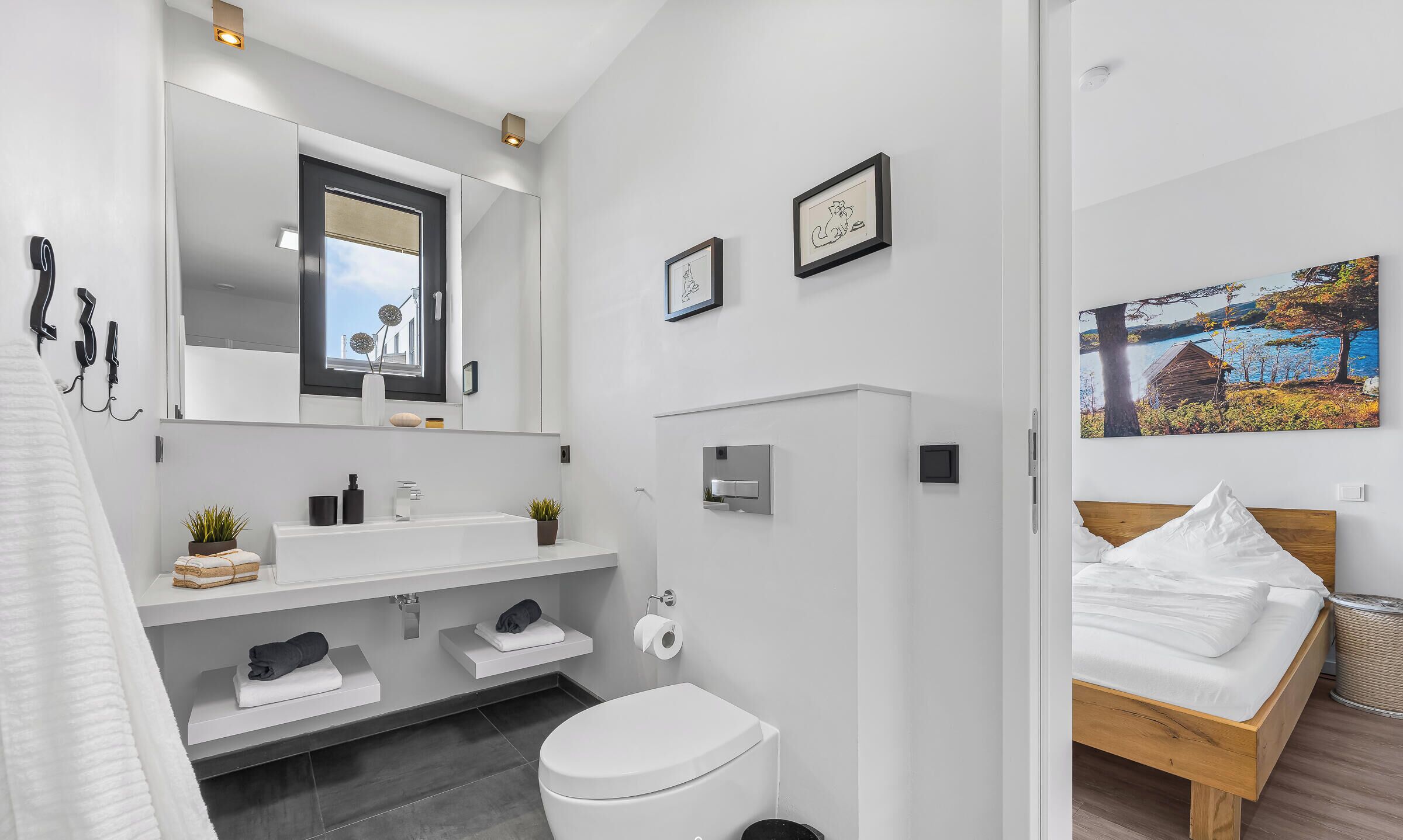 A modern bathroom with a toilet, sink, and shower. There is a bed with a wooden headboard and a painting on the wall.