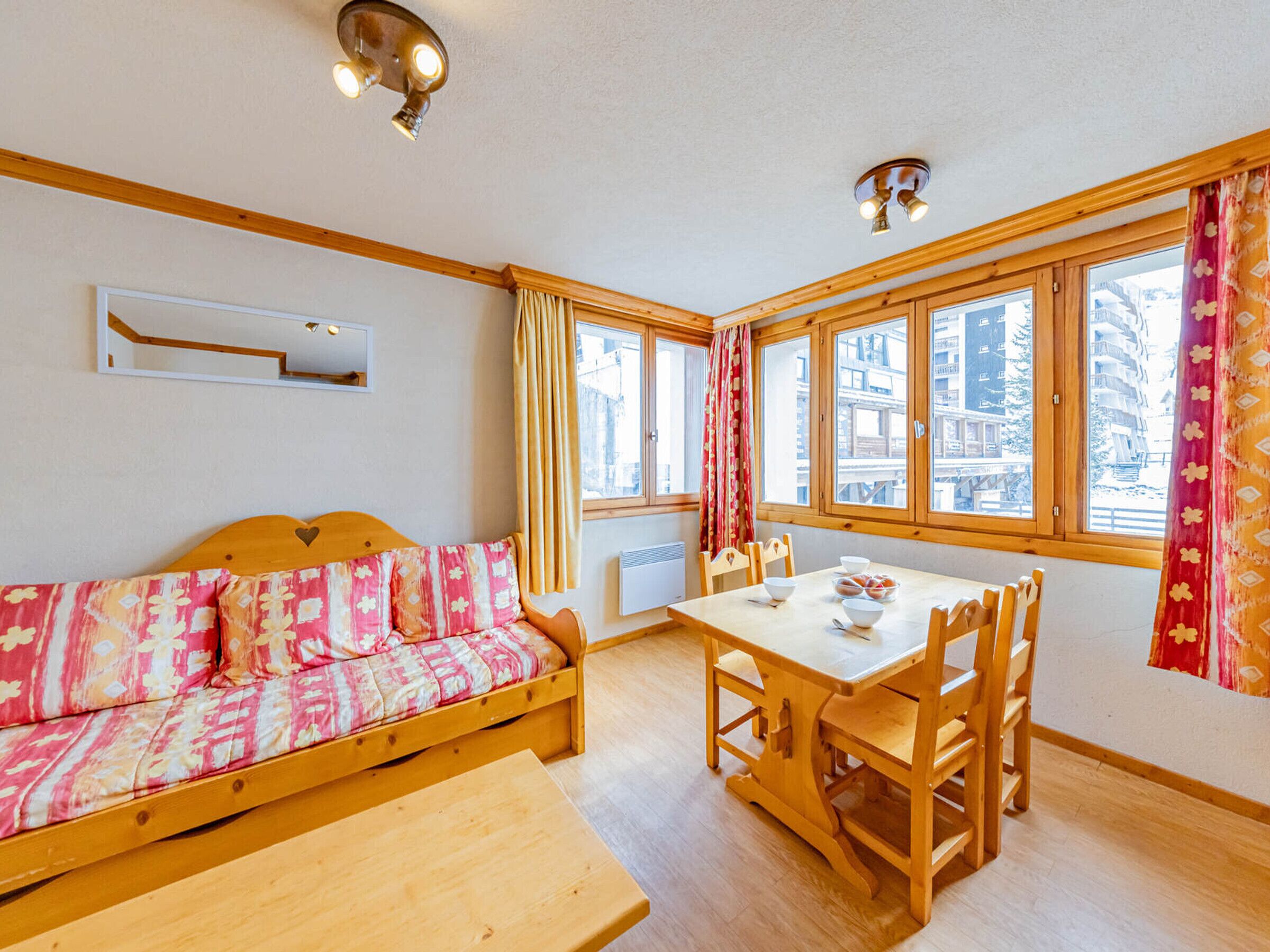 A cozy room with a wooden sofa, a dining table, and chairs. There are large windows with curtains, a ceiling light fixture, and a wall-mounted heater.