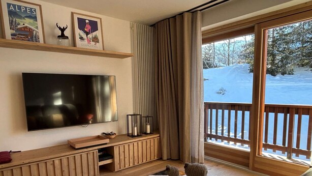 A cozy living room with a flat-screen TV, wooden furniture, and a view of a snowy landscape through large windows. - Renovated apartment in Méribel, near slopes, fiber included (Méribel)