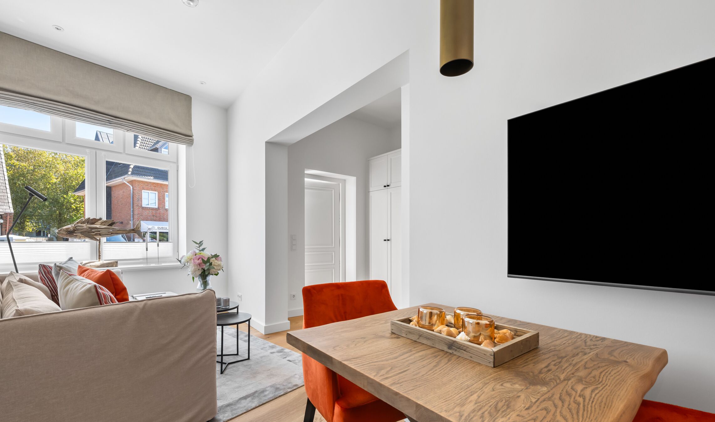 Een moderne woonkamer met een houten eettafel, oranje stoelen, een bank en een grote televisie aan de muur.