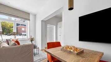 Een moderne woonkamer met een houten eettafel, oranje stoelen, een bank en een grote televisie aan de muur.