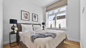 Un dormitorio con cama, dos mesitas de noche, una ventana con persianas y dos cuadros en la pared.