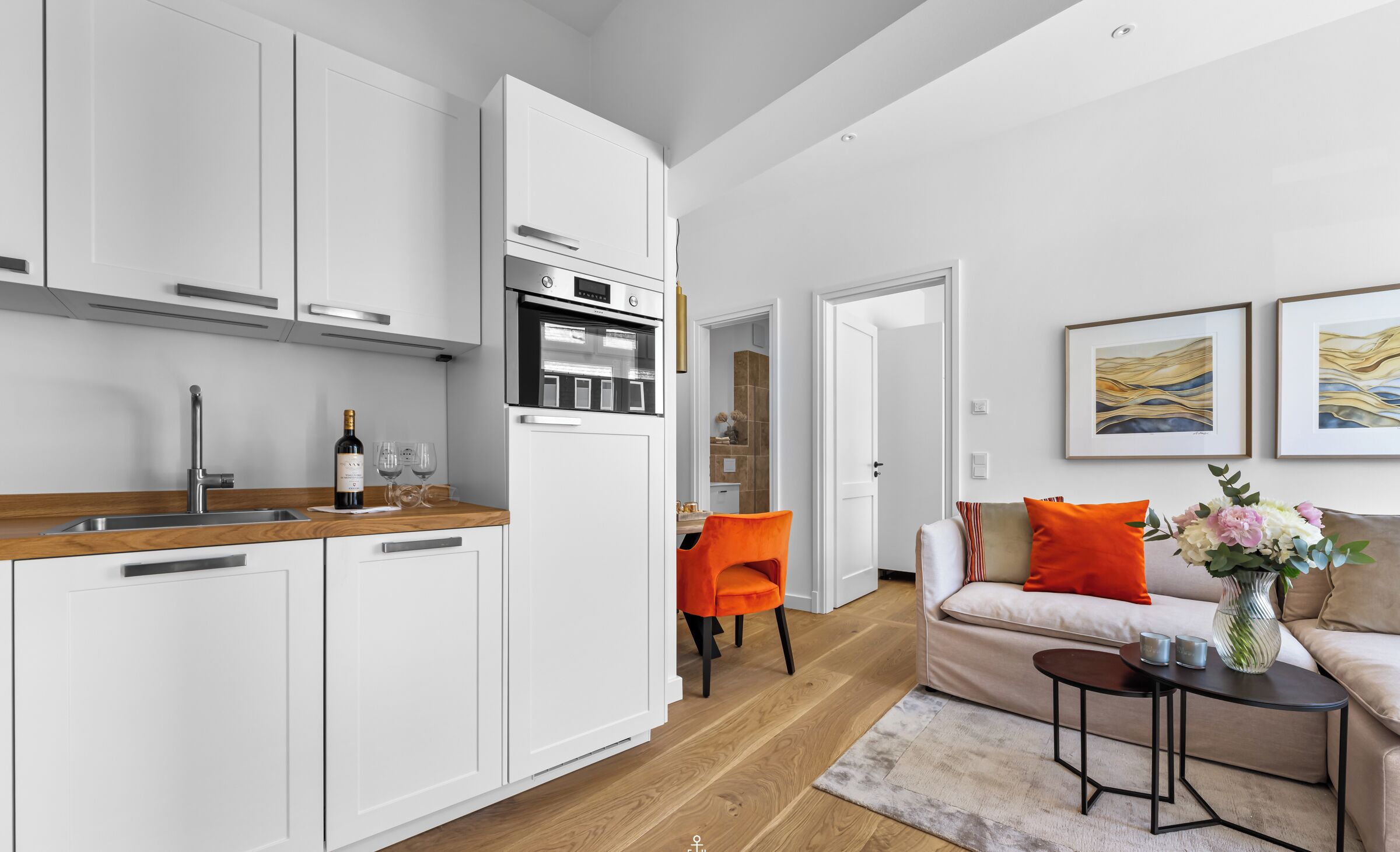 A modern kitchen with white cabinetry and a built-in oven. There is a dining area with a wooden table and orange chairs. A beige sofa and a small side table with a vase of flowers are in the living room.