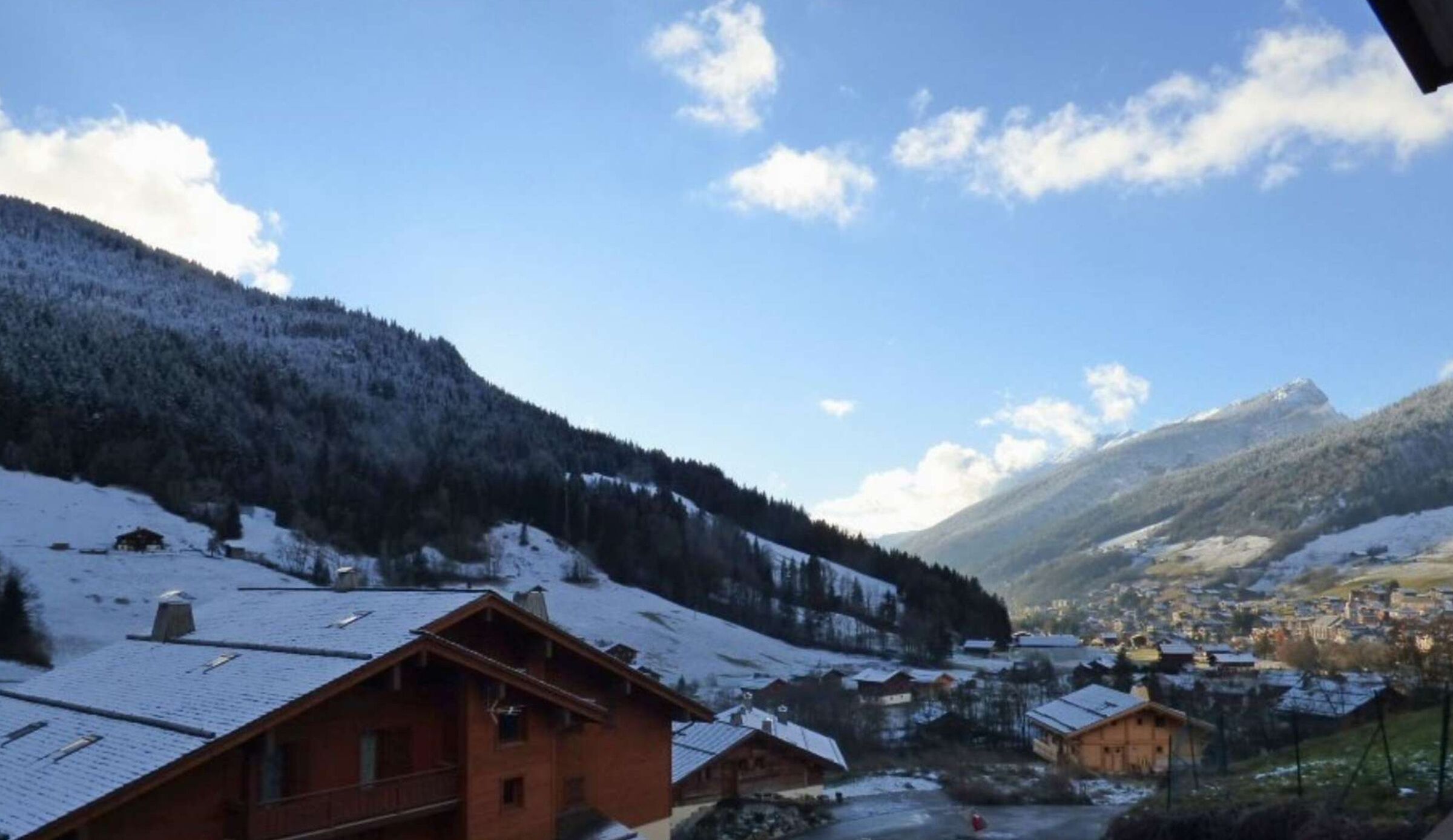 A mountain village with snow-covered roofs and buildings, surrounded by forested slopes and a clear sky.
