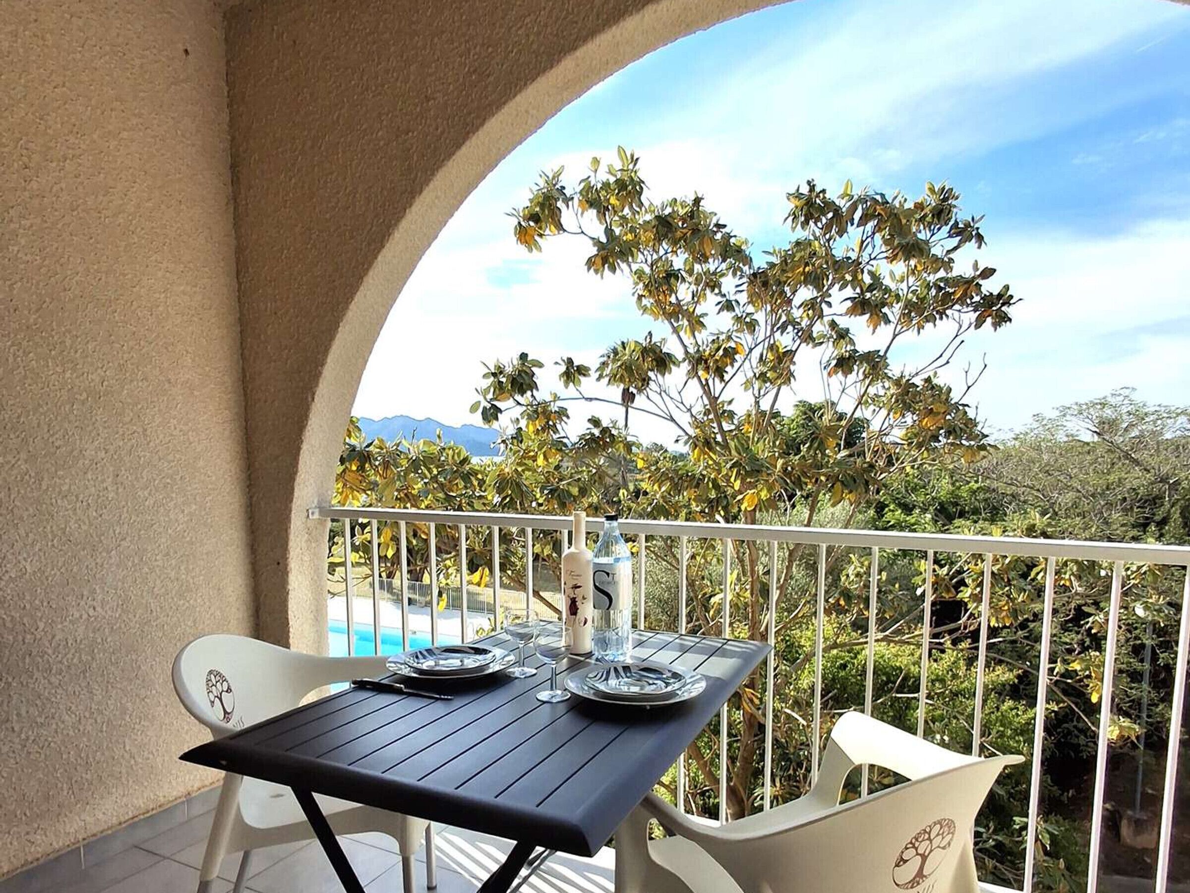 A balcony with a table set for two, overlooking a pool and trees.