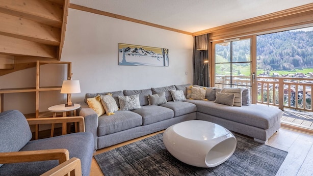 Ein modernes Wohnzimmer mit einem grauen Ecksofa, einem runden weißen Couchtisch und Blick auf eine Berglandschaft durch große Fenster.