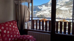 A room with a red patterned sofa, a window with a view of snow-covered mountains, and a wooden railing. - Charming ski-in T3, 6 pax, balcony, garage (Le Grand-Bornand)