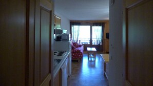 A narrow hallway leading to a kitchen with a stove and microwave, a dining area with a table and chairs, and a window with curtains. - Charming ski-in T3, 6 pax, balcony, garage (Le Grand-Bornand)