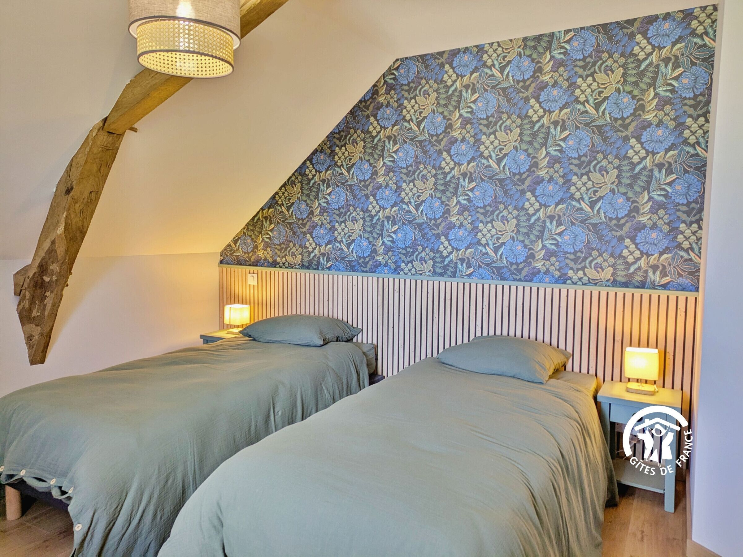 A bedroom with two beds, wooden beams, a sloped ceiling, and a floral-patterned wall.